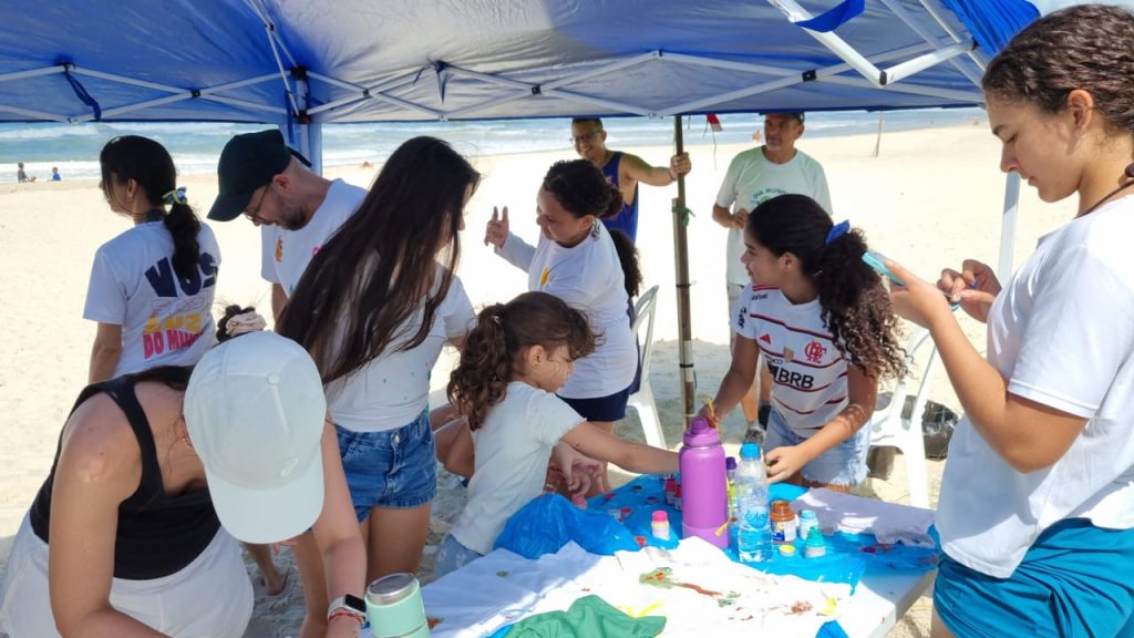 Crianças e jovens reunidos sob tenda azul realizando oficina de arte e pintura em camisetas na praia.