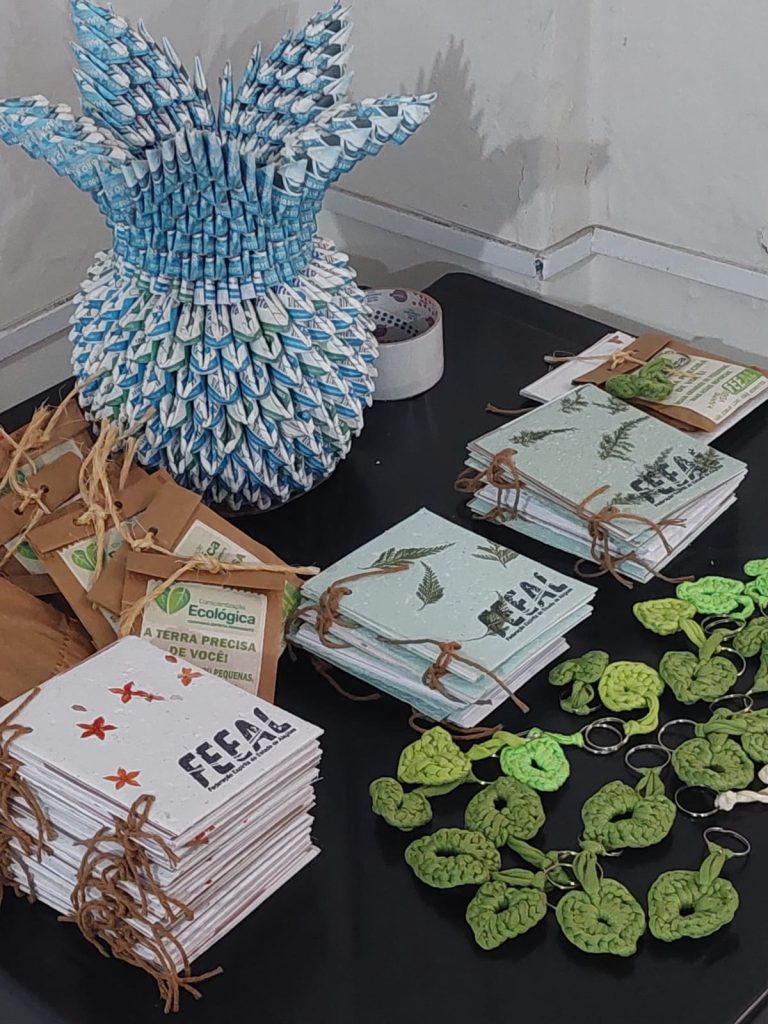 Mesa com brindes ecológicos artesanais, blocos de notas com marca da FEEAL e enfeites reciclados durante encerramento da Campanha Junho Verde.