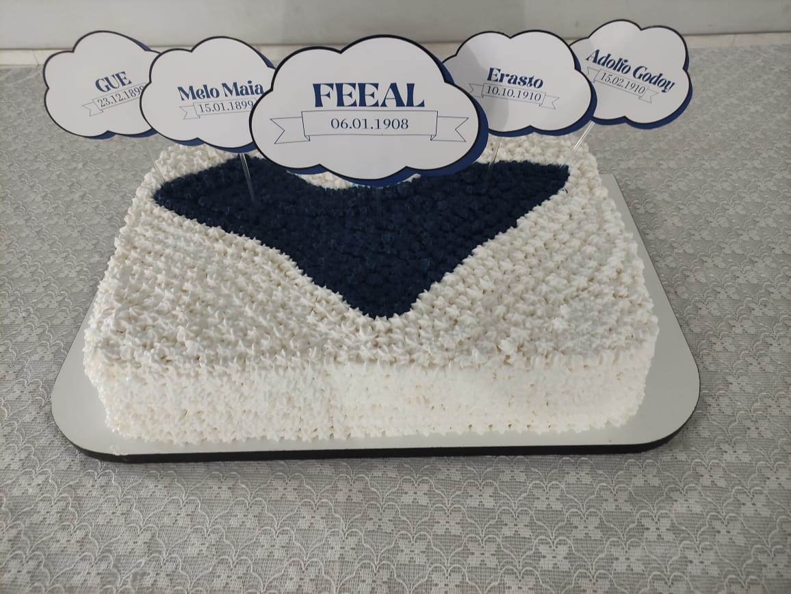 Bolo decorado em glacê branco e azul com topper central escrito FEEAL 06.01.1908 e toppers com nomes de pioneiros espíritas.